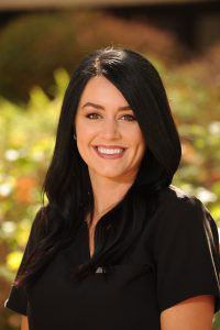 The image is a portrait of a woman with dark hair, wearing a black top and smiling at the camera. She has a professional appearance, possibly in a business or corporate setting.