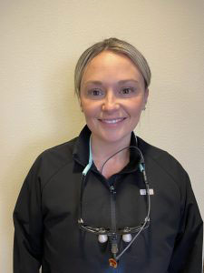 The image shows a person posing for the camera, wearing a dark jacket and a necklace with multiple beads. They have short hair and are smiling at the camera.