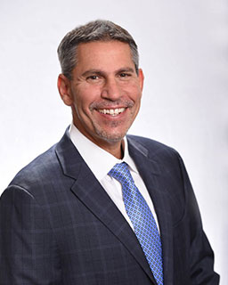 The image shows a man smiling at the camera, wearing a suit and tie. He has short hair and appears to be in a professional setting.