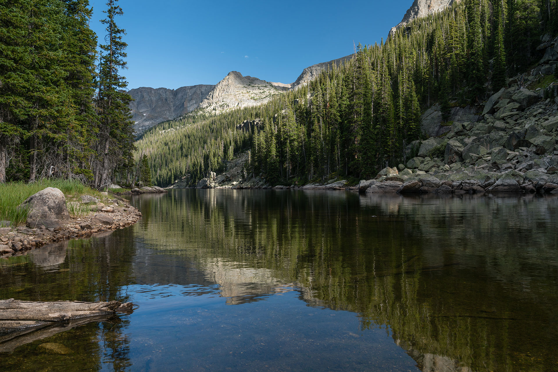 The image shows a serene mountain lake surrounded by tall trees and rugged terrain, with clear blue skies reflecting on the water.