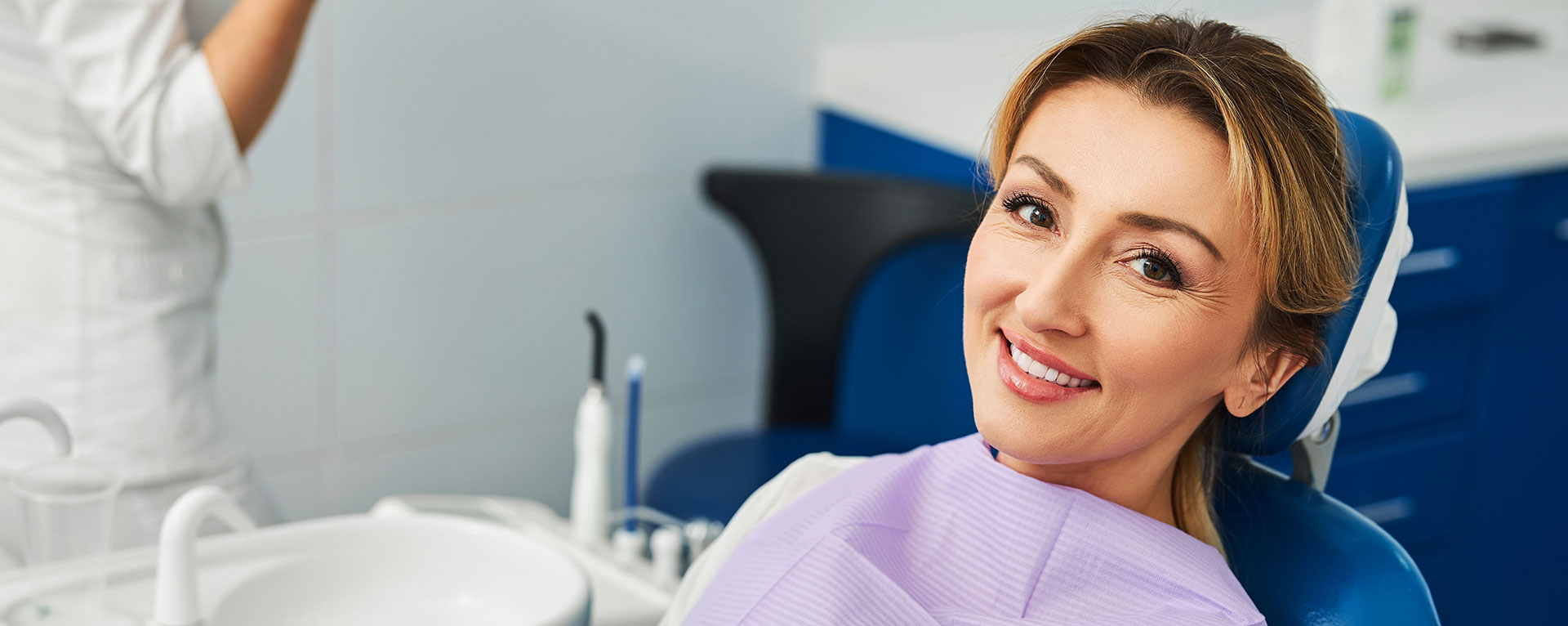 The image shows a woman in a dental office, smiling and looking directly at the camera. She is seated in a dental chair with a dental hygienist standing behind her, both wearing face masks. They are surrounded by dental equipment and tools.