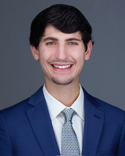 A smiling young man in a suit and tie, with a beard, against a neutral background.