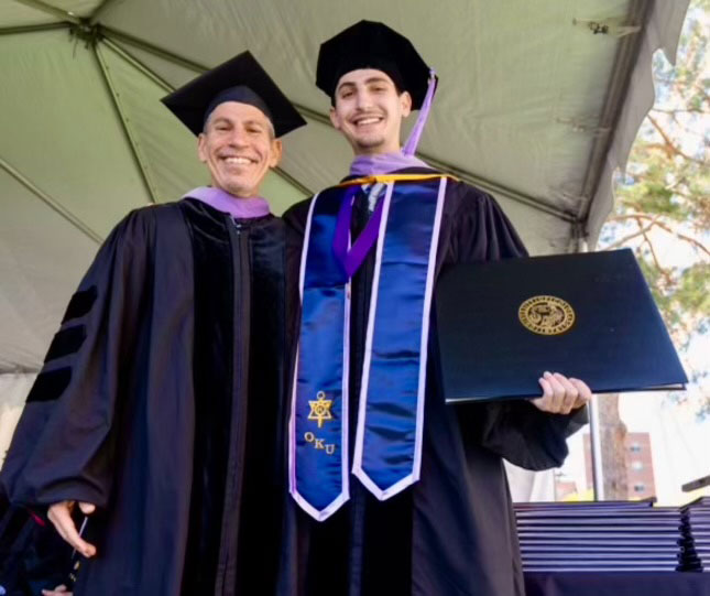 The image shows two individuals in academic regalia, one wearing a graduation cap and gown and the other holding a diploma, standing together at an outdoor event.