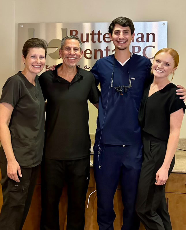 The image is a photograph of a group of four adults posing together, with one woman standing between two men and another man on the right. They are smiling and appear to be in a professional setting, possibly an office or clinic, as suggested by the signage and the attire of the individuals, which includes scrubs for the men. The environment is well-lit and the group seems to be posing for a formal occasion or event.