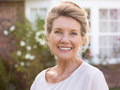 An elderly woman with blonde hair wearing a white top is standing outdoors in front of a house.