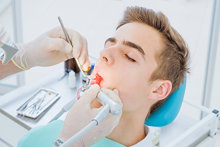 An adult male patient receiving dental care, with a dentist using a drill on his teeth while the patient is seated in a dental chair.