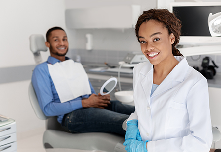 A woman in a dental office, smiling and wearing a white coat, is standing next to a man who is seated and appears to be a patient. Both are surrounded by dental equipment and devices.