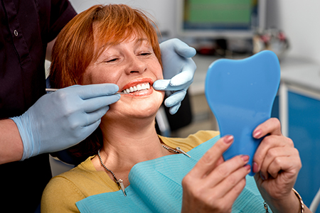 A woman in a dental chair receiving a teeth cleaning, with a dentist performing the procedure and holding up a tooth model for her inspection.