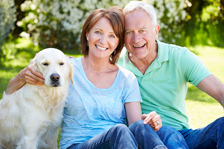 A man and woman sitting on the grass with a golden retriever between them, smiling at the camera.