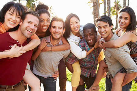 The image shows a group of young adults posing together for a photo, with one person in the center holding up their arms to embrace others. They appear to be outdoors during the daytime, with a clear sky and palm trees in the background, suggesting a warm climate.