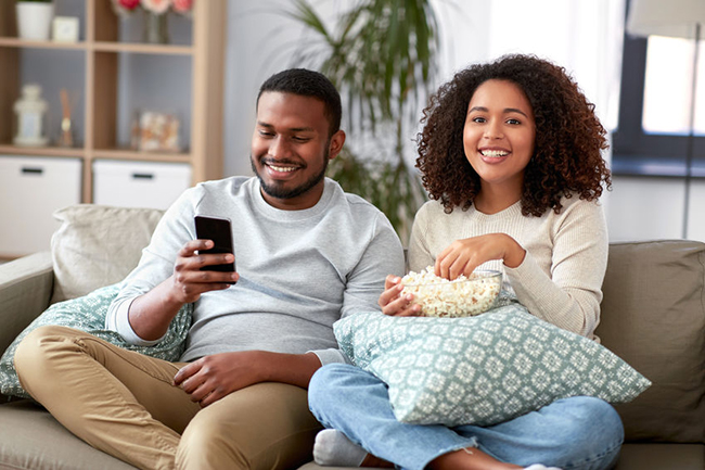 Two people, a man and a woman, are sitting on a couch with snacks, enjoying each other s company while watching something on a phone.